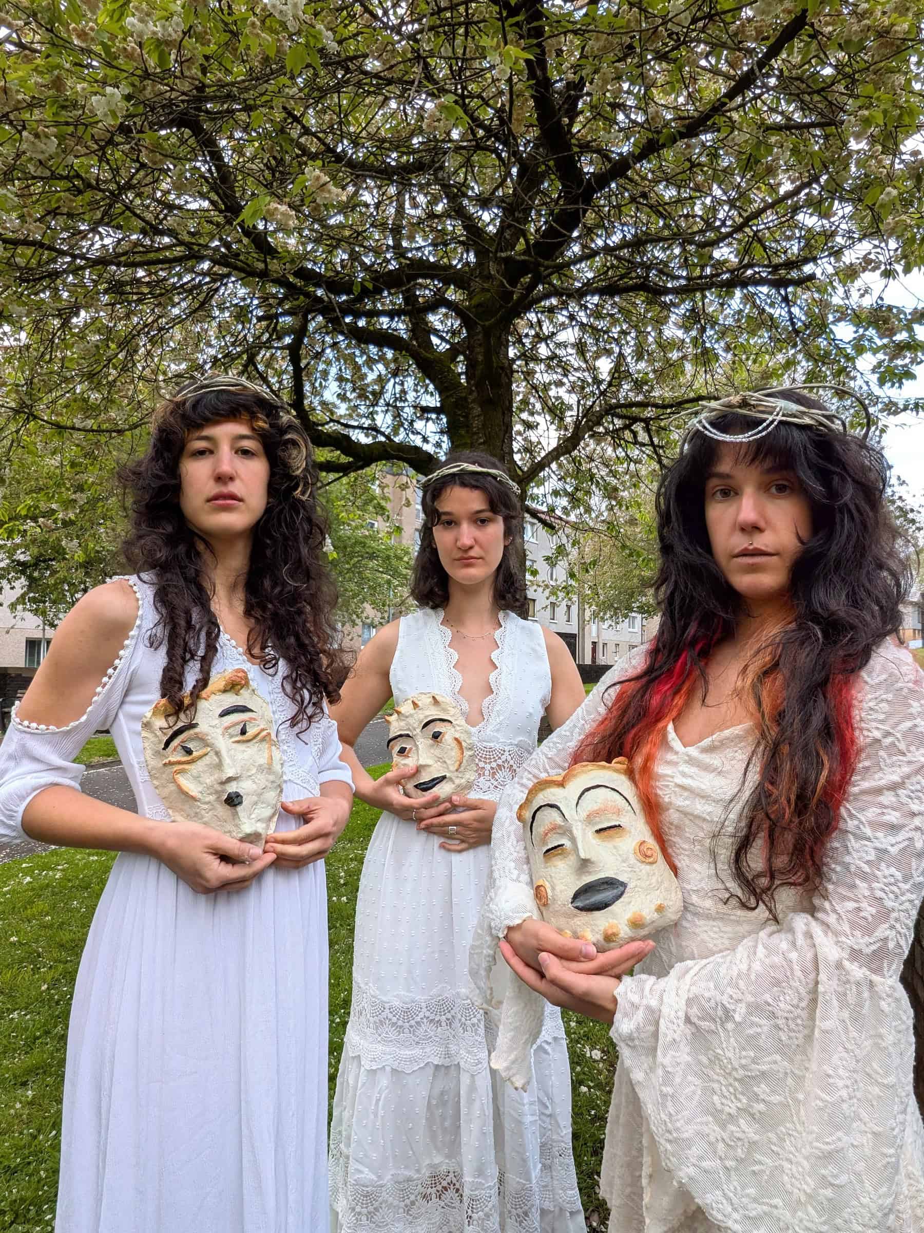 three women in white dresses stand outside holding masks, part of Terpsikores_Work in progress What the Land still Knows