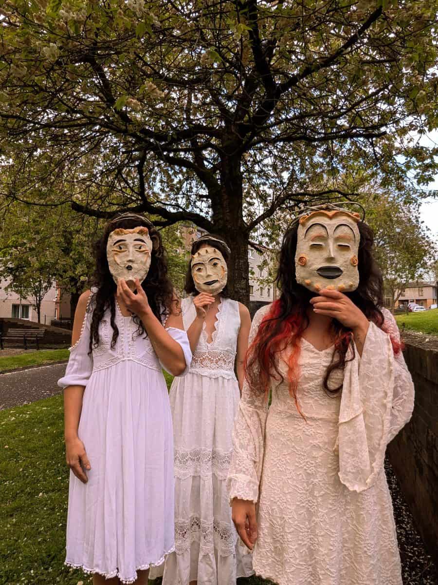 three women in white dresses stand outside wearing masks, part of Terpsikores_Work in progress What the Land still Knows